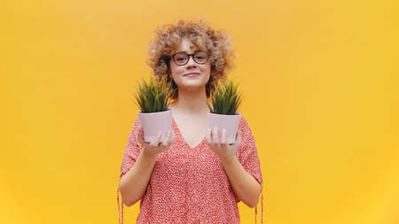 Attractive girl holding two small pot plants in her hands. Girl wearing round spectacles dressed in casual pink top with flowers in her curly hair. Isolated with yellow background studio.の写真素材