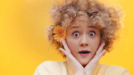 Cute surprised girl posing with orange gerbera daisy flower on her ear. Dressed in a yellow sweater. Isolated girl with bright yellow background studio. Girl looking surprised.の写真素材