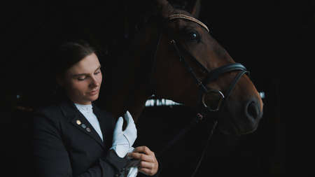Female jockey putting on her gloves. A Seal brown horse is standing by her side in the stable. Black background. Jockey getting ready for the horse riding competition.の写真素材