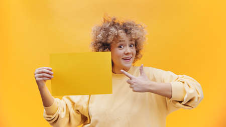 Girl holding a yellow sheet of paper pointing at the paper. Isolated girl over a bright yellow...