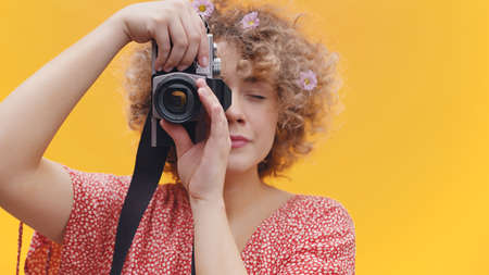 Attractive girl holding a vintage camera taking a picture. Girl in a joyful mood over bright yellow background studio. Dressed in a casual pink shirt wearing flowers in her hair. Photography concept.の写真素材