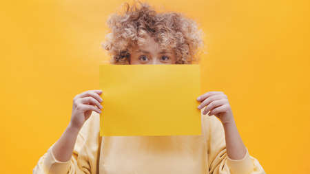 Girl holding a yellow sheet of paper covering half of her face with the paper. Isolated girl over a bright yellow background with yellow paper. The girl is dressed in a yellow baggy top.の写真素材