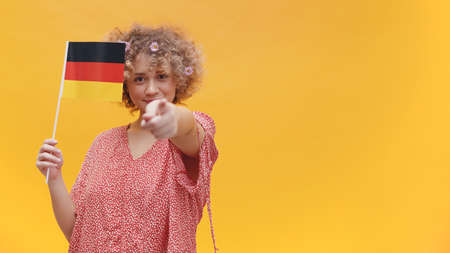 Girl holding German flag in her hand. Pointing with her finger at the camera. Girl isolated over a yellow background with Deutschland Flag. Learn German concepts. Going to Deutschland.の写真素材