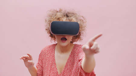 Girl wearing Virtual Reality glasses watching a 3D movie. Feeling amazed and pointing at the virtual screen. The girl is lost in virtual reality. Isolated over pink background studio.の写真素材