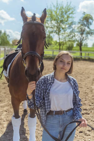 Young girl with her Bay horse standing in the sandy arena. Girl holding horse lead rope looking straight at the camera. Posing with the stallion. Sandy arena with wooden fence in the background.の写真素材