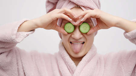 Girl with a head towel wearing a pink bathrobe holding cucumber slices in her hands. Covering her eyes with the cucumber. Concept of skincare while staying at home. Girl in a good mood pampering herself.の写真素材