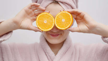 Joyful girl wearing head towel and a bathrobe holding two orange slices in her hand. Covering her eyes with the orange. Concept of taking care of skin and hydrating it.の写真素材