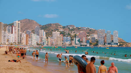 Benidorm, Spain 18.08.2021 People enjoying the beach on a sunny day. Swimming, Sunbathing, Playing and having a good time. Apartment in the background against the clear blue sky. Vacation concept.のeditorial素材