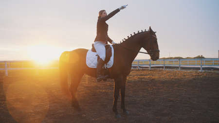 a young beautiful woman is sitting on a horse, and behind her is the sunset and she is pointing her finger at the sky. High-quality photoの写真素材