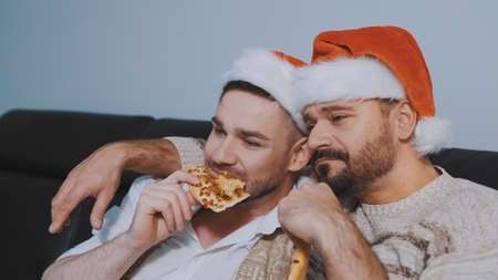 Handsome gay male couple with santa hats eating pizza and watching movie on christmas eve. High quality photoの写真素材
