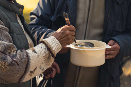 Sharing warm food with people in need concept. Cooking pot full of soup is handled between homeless people who share it with each other. High quality photoの写真素材