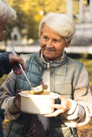 Wholesome caucasian grandmother shares her homemade soup with homeless people in park. Helping people in need. Vertical Shot. High quality photoの写真素材