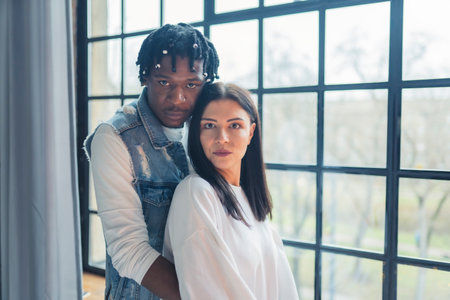 Multiracial couple standing next to the broad window, embracing, and seriously looking at camera. High quality photoの写真素材