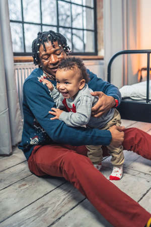 African-American Father Supporting His Toddler While Sitting On The Floor. Vertical Indoor Shot....