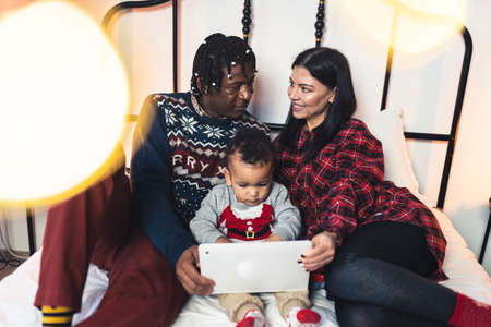 Modern family Christmas concept. African-American man and his European wife laying down on their bed with cute toddler looking at a tablet. High quality photoの写真素材