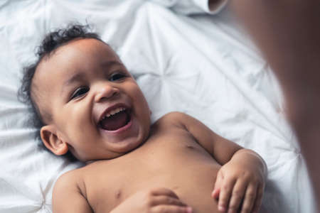 Cheerful African-American baby laying down on white sheets and smiling at his parents. High quality...