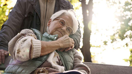 Calm elderly lady hugs her retired husbands hand while sitting on a bench in a park. High quality...
