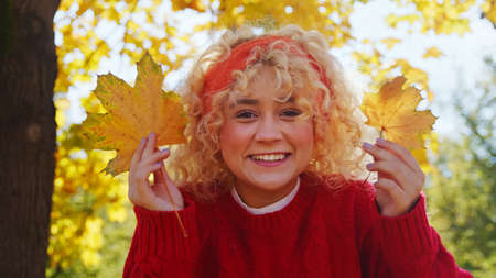 Happy young millenial girl in red with blond curly hair holding autumn leaves - portrait. High...