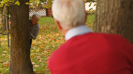 Aged Caucasian couple looking at each other from behind the trees fall man is photographed from the back, focus on the woman full shot park background . High quality photoの写真素材