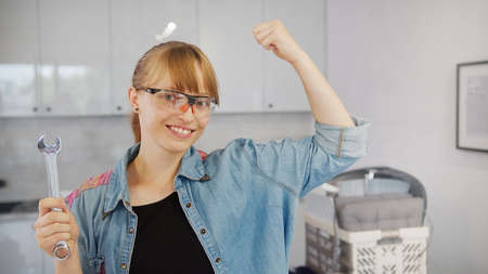 Independent caucasian woman in a jean shirt holding a wrench tool, looking at camera with one hand up, showing off her muscles. High quality photoの写真素材