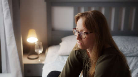 Sad depressed caucasian red-haired woman sitting down on bed in her bedroom. Anxiety concept. High quality photoの写真素材