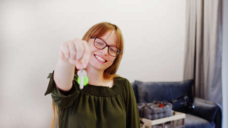 Thrilled European ginger-haired woman smiling and showing keys to the camera while standing in her living room. High quality photoの写真素材