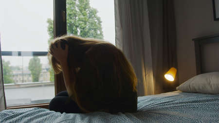 Devastated caucasian woman touching her long hair, sitting on bed at home, thinking about problems. High quality photoの写真素材