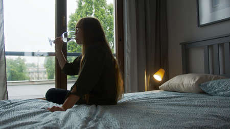 Alcohol addiction concept. Lonely sad caucasian long-haired woman drinking another wine glass while sitting on a bed. High quality photoの写真素材