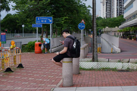 Hong Kong boy sitting and waiting on a concrete pole. Looking at and browsing on his mobile phone.のeditorial素材
