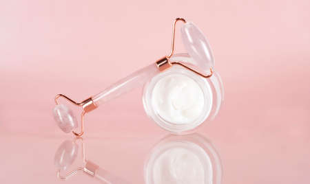 Face cream and gouache jade massage roller for face and neck massage. Lifting for healthy and youthful skin. Home facial skin care. Organic facial cosmetics on a pink background. Spa treatments.の写真素材