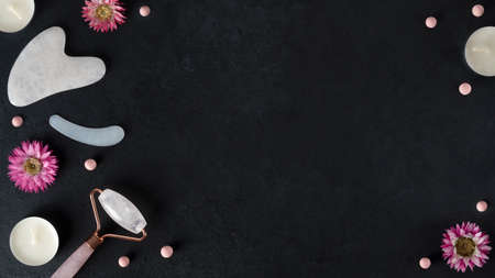 Spa treatments with gouache scraper and facial massage roller. Trending skin care. Lifting for youthful skin. Organic cosmetics on dark background with flowers and candles. Copy space. Top view.の写真素材