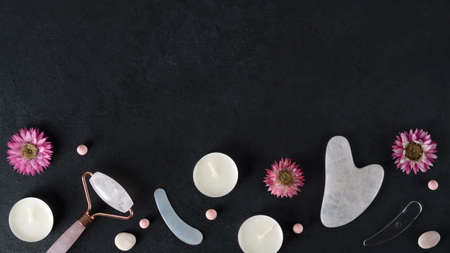 Spa treatments with gouache scraper and facial massage roller. Trending skin care. Lifting for youthful skin. Organic cosmetics on dark background with flowers and candles. Copy space. Top view.の写真素材