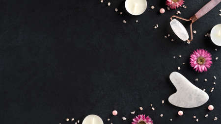 Spa treatments with gouache scraper and facial massage roller. Trending skin care. Lifting for youthful skin. Organic cosmetics on dark background with sea salt and candles. Copy space. Top view.の写真素材