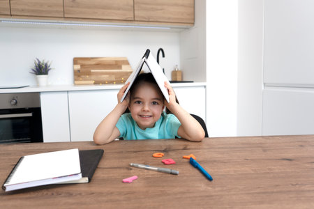 Cute smiling preschooler girl sitting at desk and holding open book above her head.の写真素材