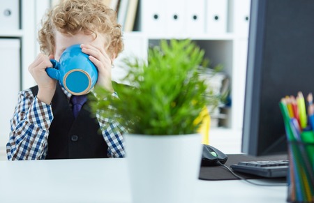 Young boy drinks from mug sitting at workplace in officeの写真素材