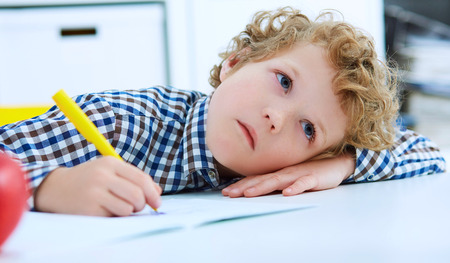 Tired little boy sitting at the table, with hishead on his arm, writing in her notebook while doing homework. Concept of a large number of assignments at school.の写真素材