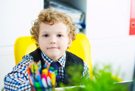 Little caucasian curly boy looking at camera sitting at computer in office. Schoolboy plays in the boss.の写真素材
