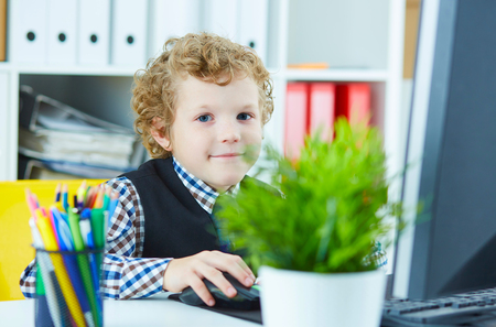 Little boy sits behind a working computer in the office looks into the camera and smiles. Boy plays in the boss.の写真素材