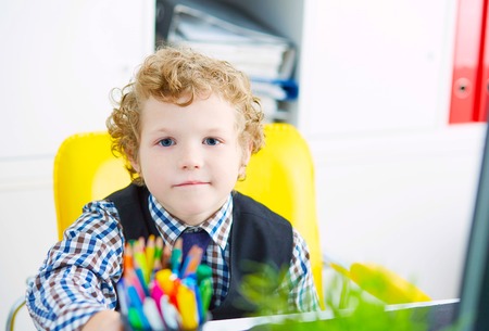 Little caucasian curly boy looking at camera sitting at computer in office. Schoolboy plays in the boss.の写真素材