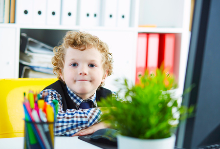 Little caucasian curly boy looking at camera sitting at computer in office. Schoolboy plays in the boss.の写真素材