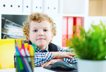 Little caucasian curly boy looking at camera sitting at computer in office. Schoolboy plays in the boss. Hands folded on a table.の写真素材