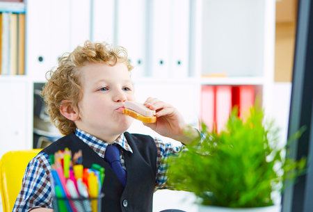 Little caucasian boy playing in the office clerk eating a sandwich with sausage in the workplace in the officeの写真素材