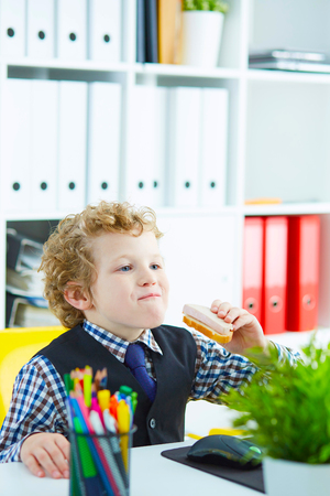 Little caucasian boy playing in the office clerk eating a sandwich with sausage in the workplace in the officeの写真素材