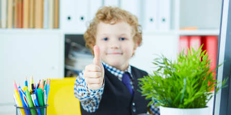 Little boy showing thumb up looking into camera and smiling. The boy is sitting in the boss's chair in the office. Selective focus on thumb.の写真素材