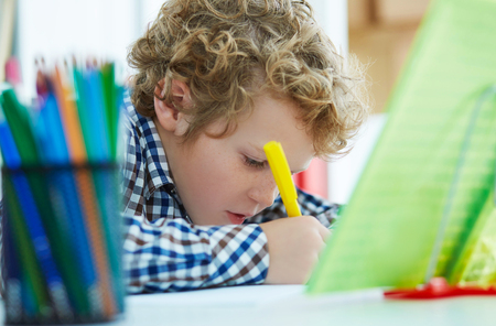 Curly Caucasian boy sitting at the desk at home and doing his homework. School, children, education conceptの写真素材