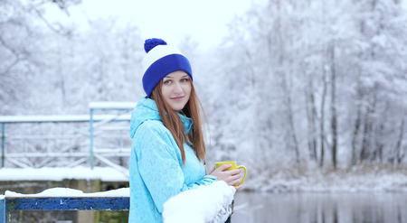Side view of young smiling woman with a cup of hot tea or coffee on snowy winter waterside.