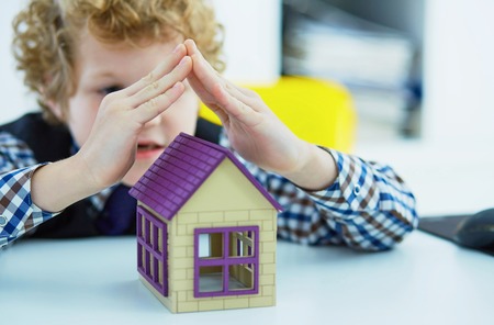 Little curly Caucasian boy holding hands over plastic figure of house at table, close up.の写真素材