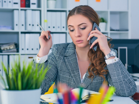 Young serious Caucasian businesswoman sitting at desk in office, speaking on the phone.の写真素材