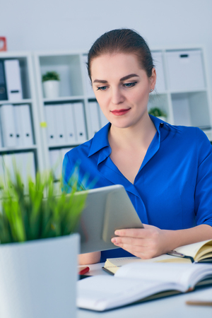 Young woman employee watching video or reading newspaper on digital tablet while sitting in modern office interior.の写真素材