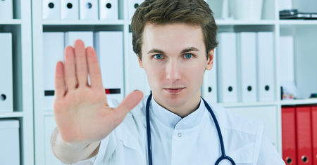 Closeup on serious male medical doctor showing stop gesture at office background. Stop gesture as he signals he has had enough, or denies access or tells someone to go away.の写真素材